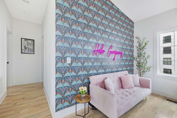 Bright modern living room corner with pastel pink velvet sofa, teal and coral art-deco wallpaper, neon 'Hello Gorgeous' sign, small round side table with flowers and a potted plant on light wood floors
