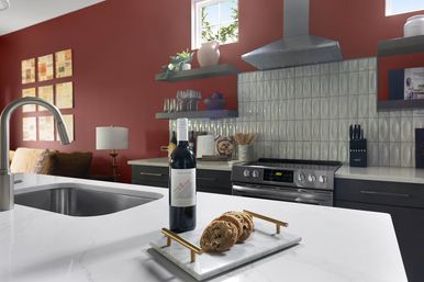 Modern kitchen with red accent wall and textured silver tile backsplash, white quartz island and stainless sink, bottle of red wine and cookies on a marble tray near a stainless gas range and open shelves.