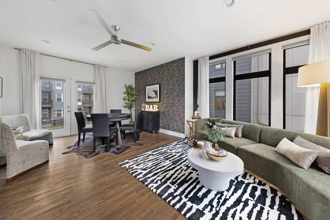Stylish bright modern urban apartment living room and dining area with curved green sectional sofa, black-and-white patterned rug, round white coffee table, dining table by balcony doors, large windows and hardwood floors.