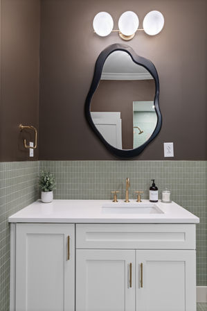 Modern bathroom vanity with white cabinets and brass hardware, sage-green mosaic tile wainscot, irregular black-framed mirror, triple globe wall light, brass faucet, soap dispenser and small potted plant.