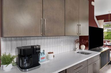 Modern kitchen countertop with black coffee maker and potted plant on a white marble surface, white subway-tile backsplash, warm wood cabinets, undermount sink with stainless faucet, decorative vase, flat-screen TV and red accent wall in an open living area.