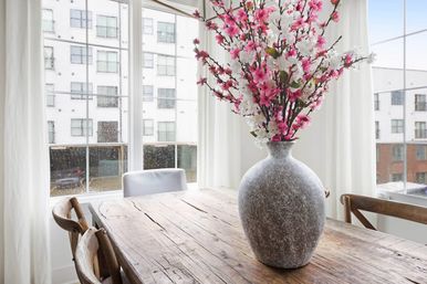 Light-filled urban dining nook with a rustic wooden table and wooden chairs, a large textured stone vase of pink and white cherry blossoms, and rain-speckled city windows in the background.