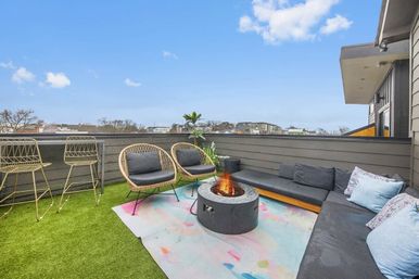 Modern urban rooftop patio with a sectional sofa, wicker chairs, bar stools and a central fire pit on a pastel rug over artificial turf, clear blue-sky skyline in the background.