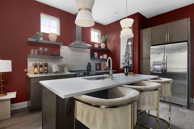 Stylish modern kitchen with deep red walls, white quartz island and breakfast bar, stainless steel refrigerator and range hood, pendant lights, open shelving and velvet-upholstered bar stools.