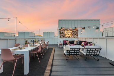Sunset urban rooftop patio with a long white dining table and pink chairs, cozy cushioned lounge, string lights and a vibrant flower wall with neon accent
