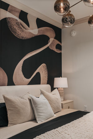 Cozy modern bedroom with neutral beige bedding and textured throw, sculptural black-and-tan abstract accent wall, bedside lamp on a nightstand and reflective glass globe pendant lights.
