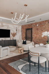 Modern open-concept urban living and dining room with exposed brick wall, sculptural white-globe chandelier, cream curved sofa and boucle chairs, wood floors, black-and-white patterned rug, media console and orchids on the dining table.