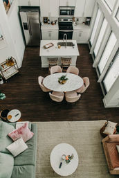 Open-concept urban loft interior with white kitchen and stainless appliances, central island, round marble dining table with blush chairs, dark hardwood floors, tall windows, and cozy pastel living area with rug.