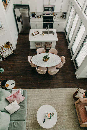 Open-concept urban loft interior with white kitchen and stainless appliances, central island, round marble dining table with blush chairs, dark hardwood floors, tall windows, and cozy pastel living area with rug.