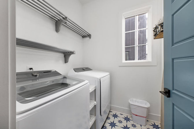 Bright, compact laundry room with top-loading washer and matching dryer, wall-mounted shelving, star-patterned blue-and-white floor tiles, small trash bin, window letting in natural light, and a teal paneled door.