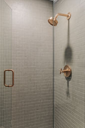 Minimalist modern shower with brushed brass showerhead and cross-handle, glass door with brass pull, surrounded by small square gray mosaic tiles.