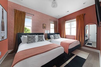 Sunlit modern bedroom with two beds, terracotta accent walls and rust curtains, black-and-white geometric pillows, textured rust bed runners, gold bedside lamp, arched full-length mirror, and wood floors.