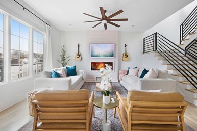 Bright modern open-concept living room with large grid windows, white sofas with teal pillows, shiplap fireplace flanked by guitars, oversized wood ceiling fan, leather armchairs, orchid centerpiece, and black‑railed staircase