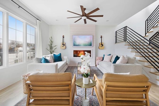 Bright modern open-concept living room with large grid windows, white sofas with teal pillows, shiplap fireplace flanked by guitars, oversized wood ceiling fan, leather armchairs, orchid centerpiece, and black‑railed staircase