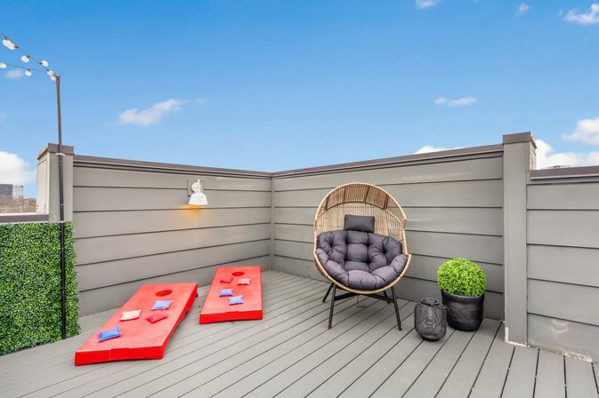 Sunny urban rooftop deck with gray privacy panels, round rattan lounge chair with plush cushion, two red cornhole boards with bean bags, string lights and potted greenery under a blue sky.