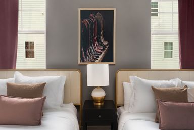 Chic twin beds in a boutique-style bedroom with beige headboards, crisp white linens and mauve throw pillows, a black nightstand with a white-and-gold lamp, framed artwork of a row of leather boots on a gray wall, and tall windows with mauve curtains letting in daylight.