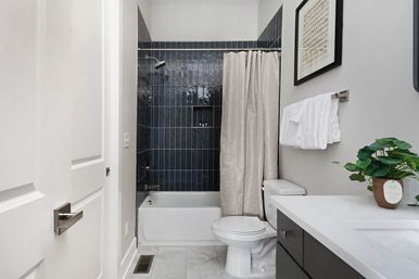 Compact modern bathroom with dark navy vertical subway-tile shower, white bathtub and toilet, gray fabric shower curtain, white countertop vanity with dark drawers, folded towels and potted plant