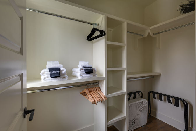 Tidy walk-in closet with white built-in shelving and chrome rods, wooden and black hangers, stacked white towels labeled 'makeup', a folding luggage rack and a box fan on hardwood floor.