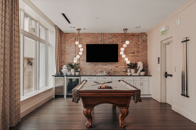 Stylish clubhouse game room with a wooden pool table, exposed brick accent wall, wall-mounted TV, sculptural pendant lights, wet bar cabinets, large windows, and hardwood floors.