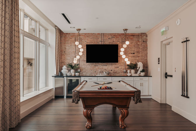 Stylish clubhouse game room with a wooden pool table, exposed brick accent wall, wall-mounted TV, sculptural pendant lights, wet bar cabinets, large windows, and hardwood floors.