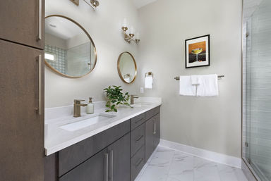 Sleek modern bathroom with double white quartz sink vanity, dark gray cabinets, two round gold mirrors, brass faucets, potted plant, wall towel rack, marble-look tile floor and glass shower