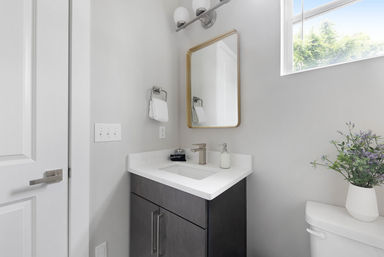 Bright minimalist modern powder room with dark wood vanity and white countertop, undermount sink with chrome faucet, gold-framed mirror and wall sconce, high window flooding natural light, towel ring and vase of purple flowers on the toilet.