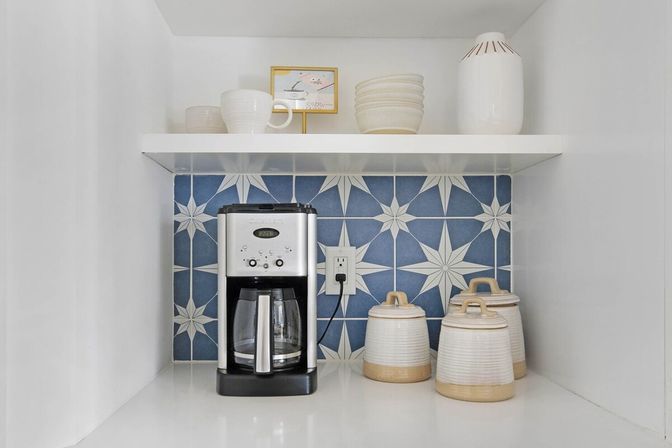 Cozy kitchen coffee station with stainless drip coffee maker, blue star-pattern tile backsplash, white floating shelf with bowls, and ceramic storage canisters