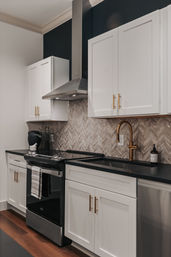 Modern kitchen with white shaker cabinets, black countertops, stainless steel range and hood, brass gooseneck faucet, chevron stone backsplash, and warm hardwood floor — sleek and cozy.