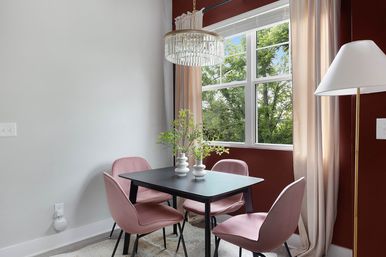 Sunlit dining nook with a black square table and four blush pink chairs, crystal chandelier overhead, large window framing green trees, maroon accent wall and cream curtains.