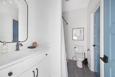 Bright modern white bathroom with a white vanity and black faucet, curved mirror, subway-tiled shower with textured curtain, toilet, gray floor tiles and a blue paneled door
