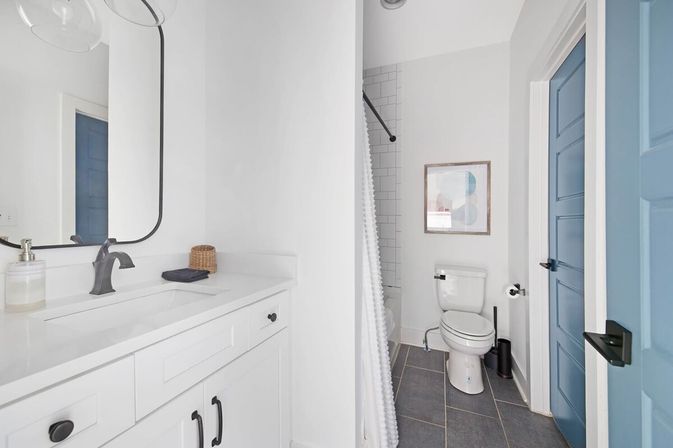 Bright modern white bathroom with a white vanity and black faucet, curved mirror, subway-tiled shower with textured curtain, toilet, gray floor tiles and a blue paneled door