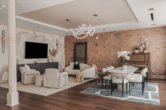 Cozy open-concept urban loft living and dining area with exposed brick wall, warm wood floors, neutral plush seating, sculptural globe chandelier, wall-mounted TV, dining table with upholstered chairs and a piano topped with plants.