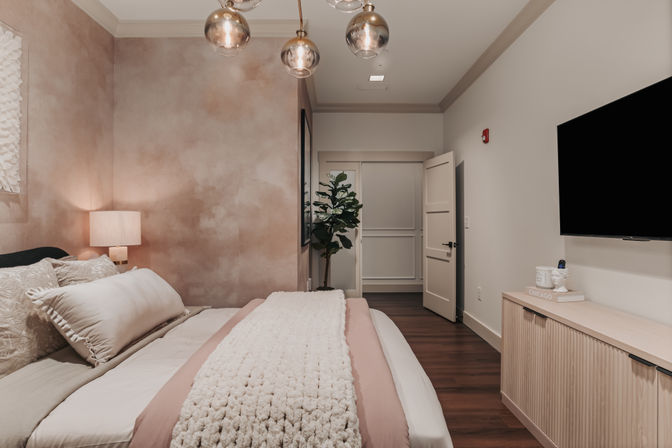 Cozy modern bedroom with soft pink textured accent wall, layered neutral bedding and chunky knit throw, wood floors, wall-mounted TV, and potted plant.