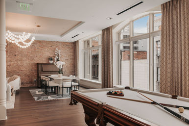 Sunlit urban loft game room with white-felt pool table and cues in foreground, round dining table and chairs on a zebra rug, exposed brick wall with piano, and large arched windows overlooking city buildings.