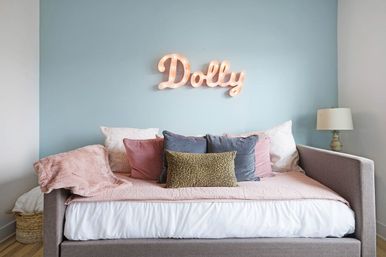 Cozy pastel bedroom with gray upholstered daybed, pink quilt and velvet pillows, blue accent wall and lit marquee sign reading "Dolly", bedside lamp and wicker basket