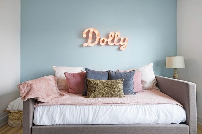 Cozy pastel bedroom with gray upholstered daybed, pink quilt and velvet pillows, blue accent wall and lit marquee sign reading "Dolly", bedside lamp and wicker basket