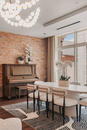Stylish sunlit urban loft dining room with an exposed brick wall, upright piano, white oval table surrounded by boucle chairs, black-and-white geometric rug, orchids centerpiece, and a cascading globe chandelier over large windows.
