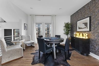Modern urban apartment game room with round poker table and four black chairs on a cowhide rug, floor-to-ceiling curtains and balcony doors showing a city view, textured accent wall with framed art and illuminated marquee "BAR" sign on a curved black cabinet.