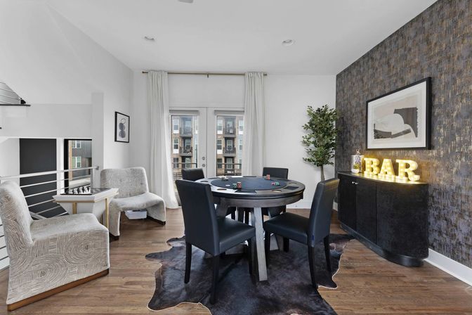 Modern urban apartment game room with round poker table and four black chairs on a cowhide rug, floor-to-ceiling curtains and balcony doors showing a city view, textured accent wall with framed art and illuminated marquee "BAR" sign on a curved black cabinet.