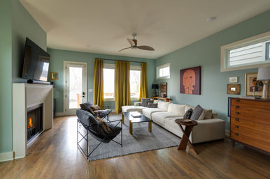 Bright, cozy modern living room with teal walls, mustard curtains, white sectional sofa, fireplace, hardwood floors, ceiling fan, abstract wall art and midcentury wooden dresser