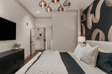 Contemporary bedroom interior with king bed, textured white throw and layered pillows, black-and-beige abstract mural wall, brass globe pendant lights, bedside lamp, flat-screen TV and dark wood media console on hardwood floor.