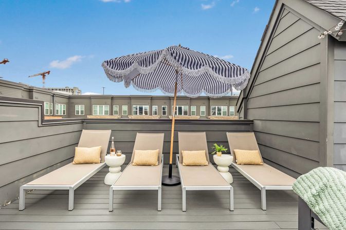Urban rooftop deck with four beige sun loungers and yellow pillows beneath a navy-and-white fringed umbrella, round side tables with drinks and a small potted plant, gray siding and city buildings beyond.