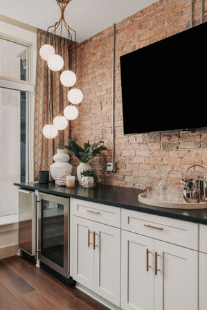 Industrial-chic kitchen bar with exposed brick wall, cascading globe pendant lights, black countertop over white shaker cabinets, built-in wine fridge, potted plants and a tray of glassware.