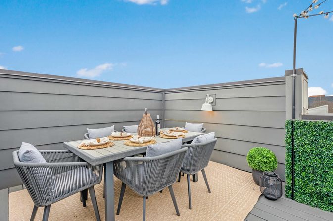 Modern rooftop patio dining on an urban terrace — gray wood-paneled walls, six-seat outdoor table with woven gray chairs and cushions, rattan lantern centerpiece, place settings, potted greenery and string lights under a blue sky.