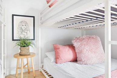 Cozy bright bedroom with a white bunk bed, soft pink quilted and fluffy pillows on the lower bed, framed line-art on the wall and a wooden stool holding a potted green plant on light hardwood floor.