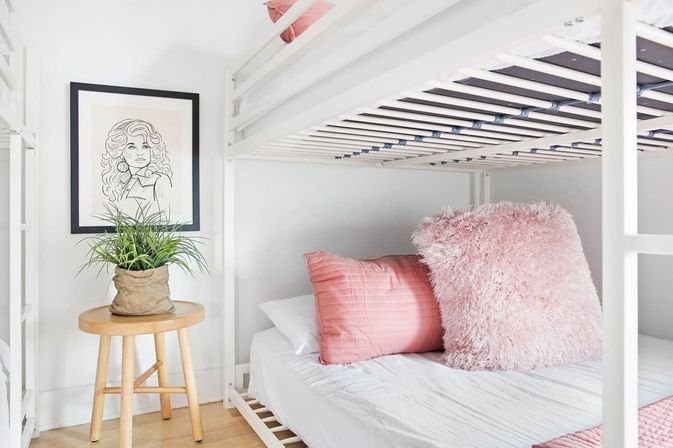 Cozy bright bedroom with a white bunk bed, soft pink quilted and fluffy pillows on the lower bed, framed line-art on the wall and a wooden stool holding a potted green plant on light hardwood floor.