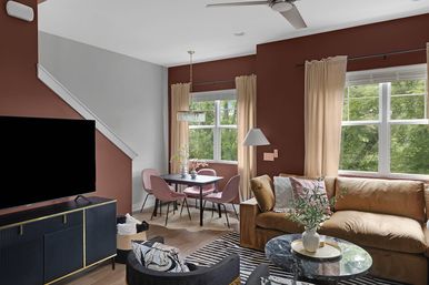 Cozy modern living room with dining nook, rust accent walls and large windows, tan velvet sofa, black marble coffee table with potted plant, round table with pink chairs under a pendant light, TV on navy console and ceiling fan