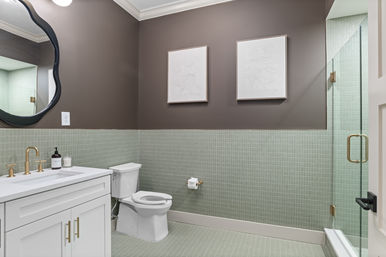 Contemporary home bathroom interior with mint-green mosaic tiles, white vanity, brass fixtures, glass shower and toilet under warm taupe walls