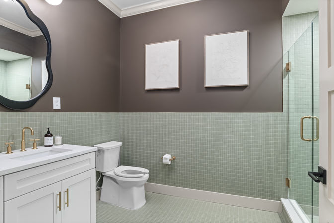 Contemporary home bathroom interior with mint-green mosaic tiles, white vanity, brass fixtures, glass shower and toilet under warm taupe walls