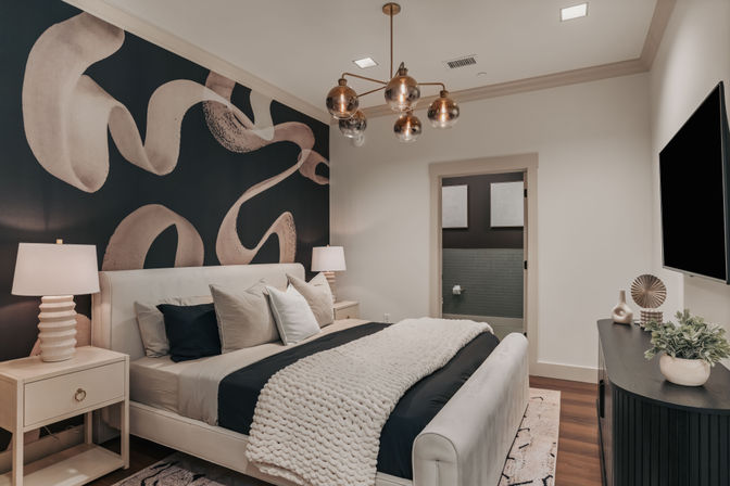 Modern master bedroom interior with abstract black-and-beige accent wall, upholstered bed with layered pillows, chunky knit throw, brass globe chandelier, bedside lamps and wall-mounted TV